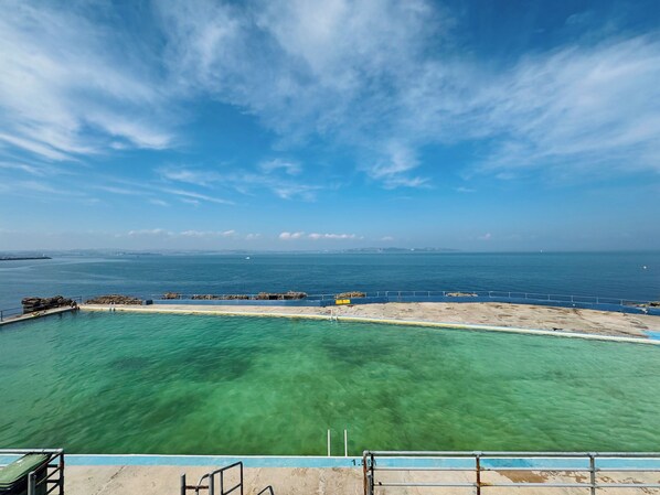 Ein klarer Blick auf einen Swimmingpool mit blauer Umrandung, einer Beton-Terrasse und eine ferne Aussicht auf das Meer und den Himmel.