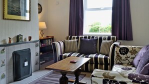 A living room with a sofa, a coffee table, a fireplace, a bookshelf, and a window with curtains. - 2 bedroom accommodation in Portinscale, near Keswick (near Portinscale)