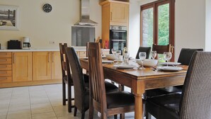 Un comedor con una mesa de madera dispuesta para cuatro personas, rodeada de sillas. Hay una zona de cocina con armarios de madera y una campana extractora de acero inoxidable.