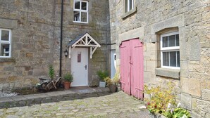 A stone building with a white door, a red door, and a small porch with a triangular roof. There are potted plants and a bicycle near the door. - 2 bedroom accommodation in Hulme End, near Hartington (Hulme End, near Hartington)