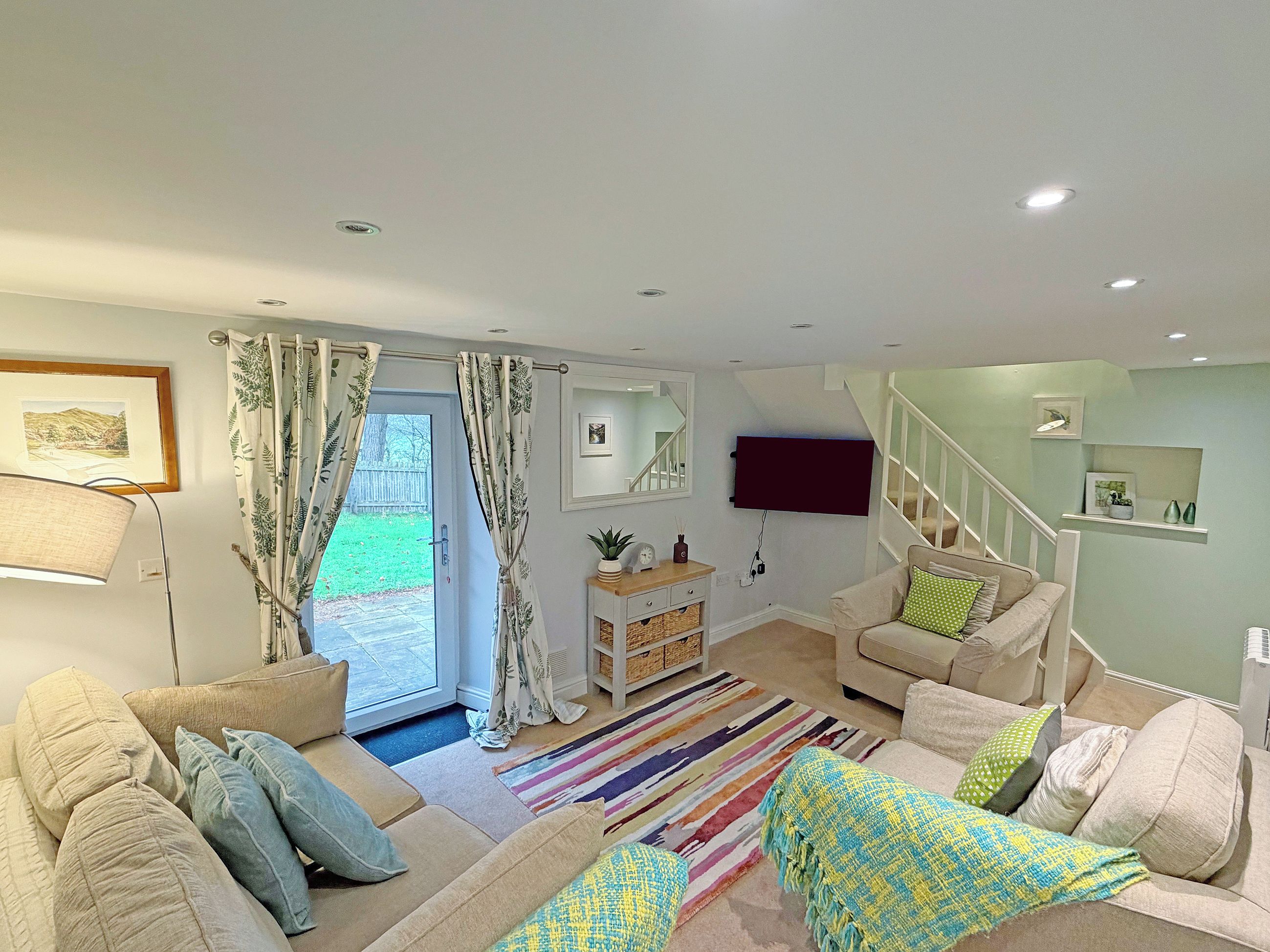 A living room with a sofa, a coffee table, a TV mounted on the wall, a staircase, and a door leading to a garden.