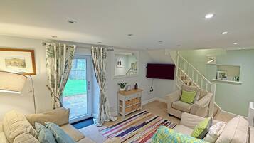 A living room with a sofa, a coffee table, a TV mounted on the wall, a staircase, and a door leading to a garden.