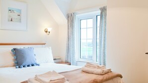 A neatly made bed with a blue pillow and a beige blanket, a small bedside table with a lamp, and a window with curtains. - 2 bedroom accommodation in Harlyn Bay, near Padstow (Harlyn Bay, near Padstow)