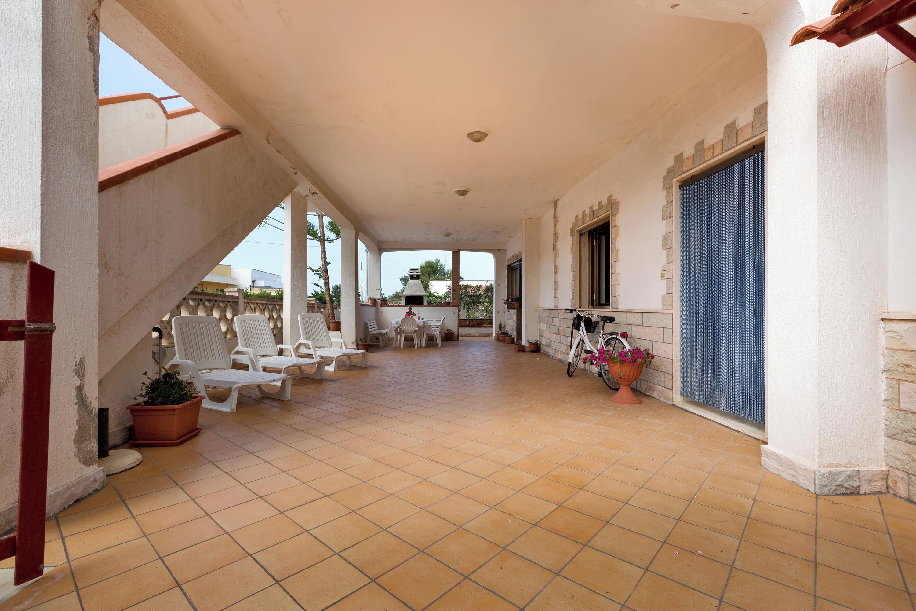 A covered outdoor area with tiled flooring, white lounge chairs, a table, and a bicycle.