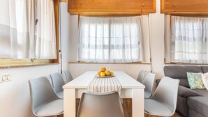 Un coin repas avec une table blanche, des chaises grises et un saladier rempli de fruits.