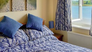 Een slaapkamer met een bed met een deken met een bladmotief, een nachtkastje met een lamp en een raam met uitzicht op een brug en water.