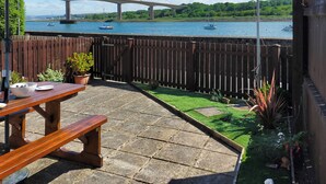 Een terras met een houten tafel en bankje, een plant in pot en een uitzicht op een rivier met boten en een brug op de achtergrond.