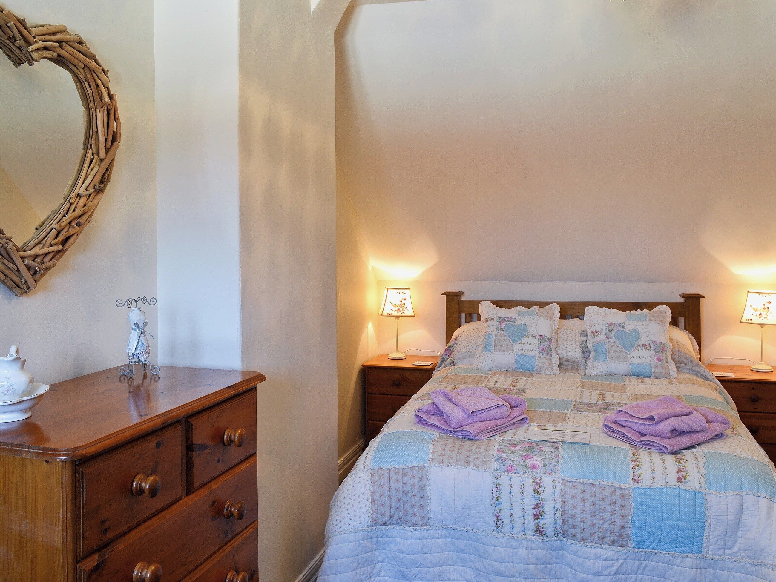 A bedroom with a wooden dresser, a mirror, a quilted bedspread, and two bedside lamps.