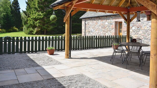 Un espacio de patio cubierto con paredes de piedra, vigas de madera y mobiliario exterior.