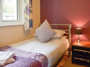 A neatly made bed with a white and purple bedspread, a single pillow, and a bedside table with a lamp.
