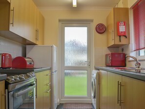 A compact kitchen with a refrigerator, microwave, and stove. There's a fire blanket on the wall and a clock above the door.