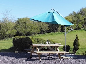 Ein Picknicktisch mit einem blauen Sonnenschirm, umgeben von Kies und Grünpflanzen.