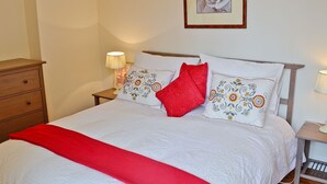 Un lit bien fait, avec du linge de lit blanc et un plaid rouge, une table de chevet en bois avec une lampe, et un tableau encadré accroché au mur.