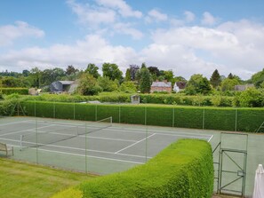 Una cancha de tenis rodeada de setos verdes y árboles, con casas al fondo.