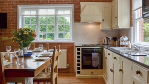 A kitchen with a brick wall, white cabinets, a dining table with chairs, and a window with a view of greenery. - 2 bedroom accommodation in Eardisland, near Leominster (Eardisland, near Leominster)