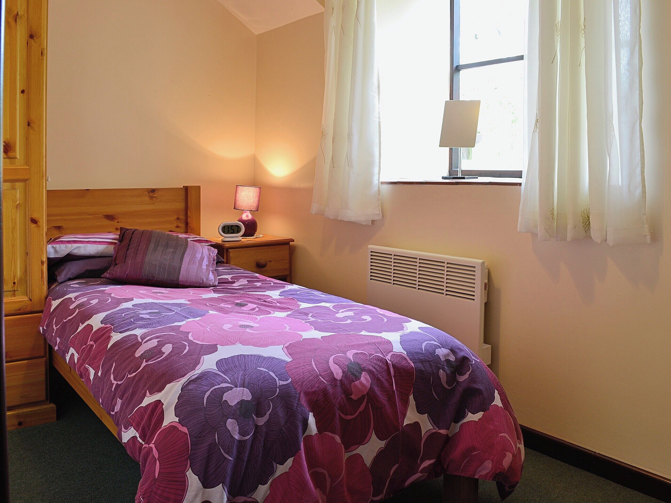 A bedroom with a bed, a nightstand with a lamp, a window with curtains, and a radiator.