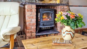 A brick fireplace with a log fire, a white leather chair, a wooden table with a 'Welcome' sign, and a vase of flowers. - Y Dderwen at Brynglas Farm (Llanfair Caereinion)