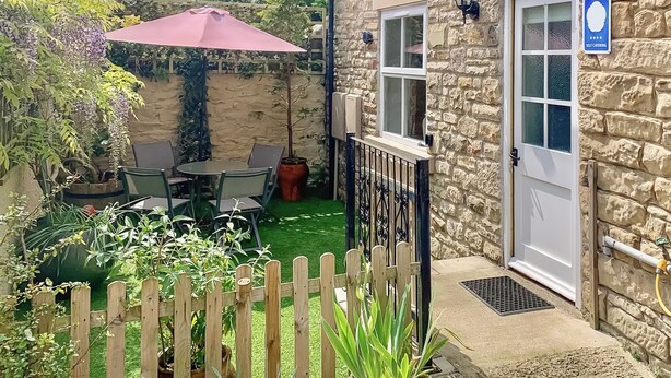 A stone building with a self-catering sign, a wooden fence, a table with chairs, and a potted plant.