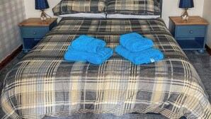 Un lit bien fait, recouvert d’une couverture à carreaux, avec deux serviettes bleues pliées posées sur le dessus. Il y a deux tables de chevet avec des lampes, et une petite table de nuit avec une lampe.