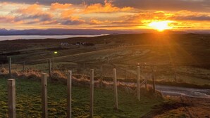 Un paysage rural avec un coucher de soleil sur une étendue d’eau, des champs et quelques bâtiments.