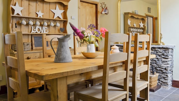 A wooden dining table with chairs, a pitcher, and a vase with flowers. There is a shelf with mugs and a sign that says "LOVE". A mirror is on the wall, and a stone wall is in the background. - 3 bedroom accommodation in Windermere (Windermere)