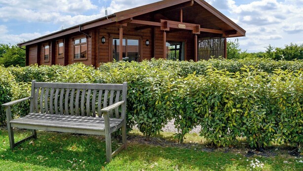 A wooden cabin with a sloped roof, a bench in front, and a well-maintained hedge. - 2 bedroom accommodation in Burgh le Marsh, near Skegness (Burgh le Marsh, near Skegness)