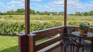 A covered outdoor seating area with a wooden bench and metal chairs, overlooking a grassy field and a body of water. - 2 bedroom accommodation in Burgh le Marsh, near Skegness (Burgh le Marsh, near Skegness)