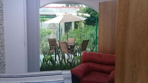 Quarto com sofá vermelho, mesa de jantar com cadeiras e vista para um jardim com guarda-sol.