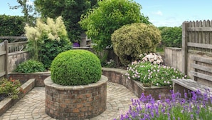 A garden with a central brick planter, surrounded by brick pathways, various plants, and a wooden fence. - 1 bedroom accommodation in Bradworthy, near Bude (Bradworthy, near Bude)