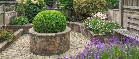 A garden with a central brick planter, surrounded by brick pathways, various plants, and a wooden fence.