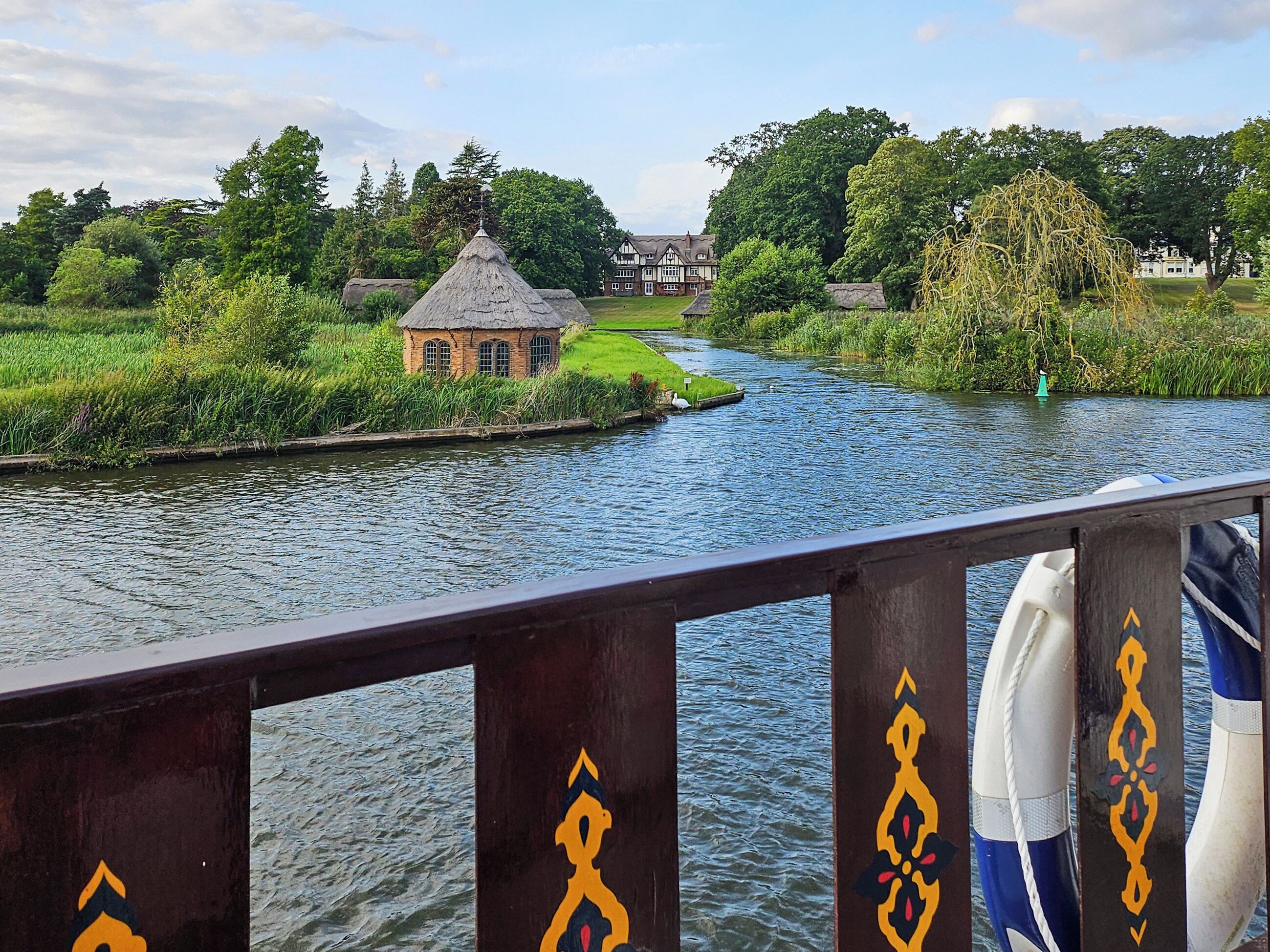 A river with a small house on the bank, a bridge railing, and a boat in the foreground.