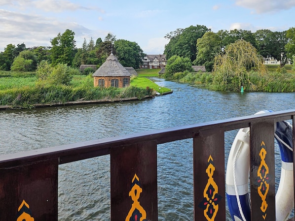 A river with a small house on the bank, a bridge railing, and a boat in the foreground.