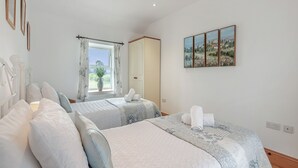 Un dormitorio con dos camas, una ventana con cortinas, un florero con una planta y un cuadro en la pared.