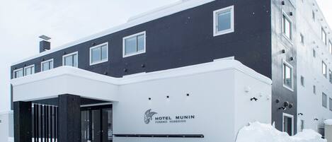 En moderne hotelbygning med mørk facade og hvide detaljer, med en markant indgang og et skilt, der viser "HOTEL MUNIN FURANO HOKKAIDO".