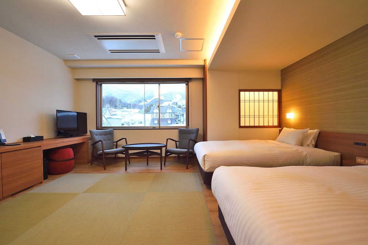A hotel room with two beds, a desk with a TV, a chair, and a view of a mountain.