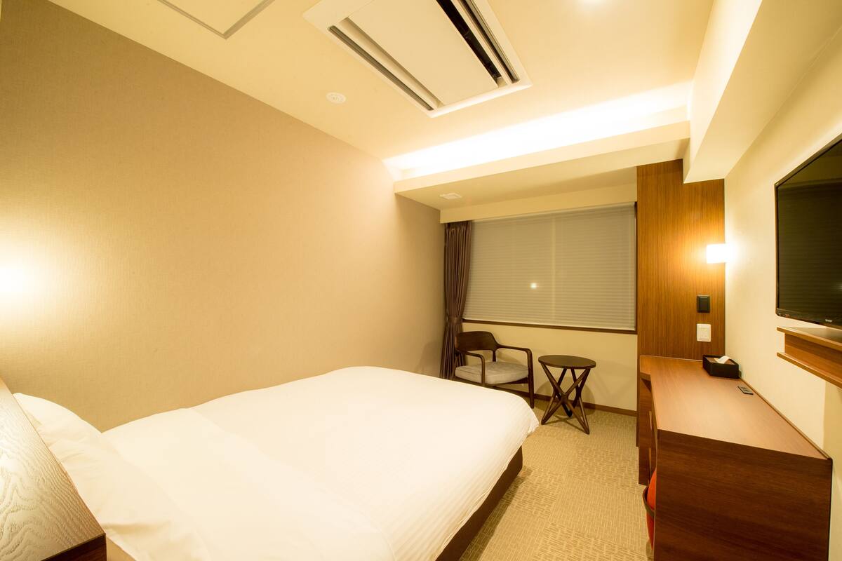 A hotel room with a large bed, a desk, a chair, a small table, a TV, and a window with curtains.