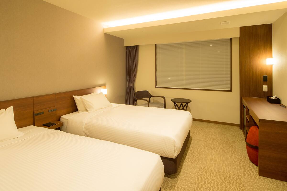 A hotel room with two beds, a desk, a chair, a small table, and a window with blinds.