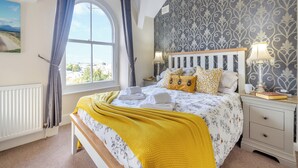 Una camera da letto con una grande finestra, un letto con lenzuola giallo e bianco, un comodino con una lampada e carta da parati decorata.