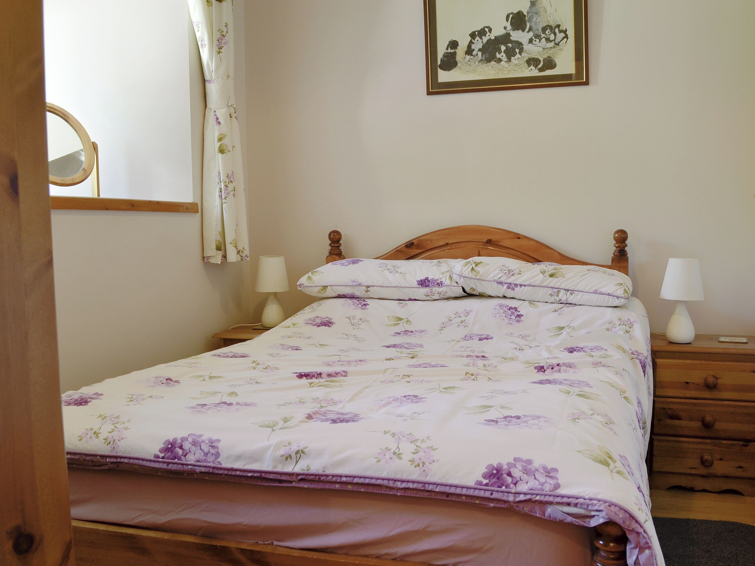 Een netjes opgemaakt bed met bloemenbeddengoed, een houten hoofdbord, twee nachtkastjes met lampen, een spiegel en een ingelijste foto aan de muur.