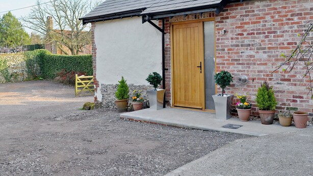Un bâtiment en briques avec une porte en bois, une allée gravillonnée et des plantes en pot.