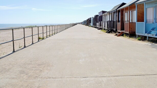 A concrete walkway along a beach with wooden beach huts on one side.