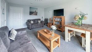 A living room with a grey sofa, a wooden coffee table, a bookshelf, and a dining table.