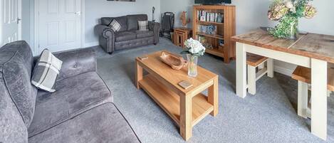 A living room with a grey sofa, a wooden coffee table, a bookshelf, and a dining table.
