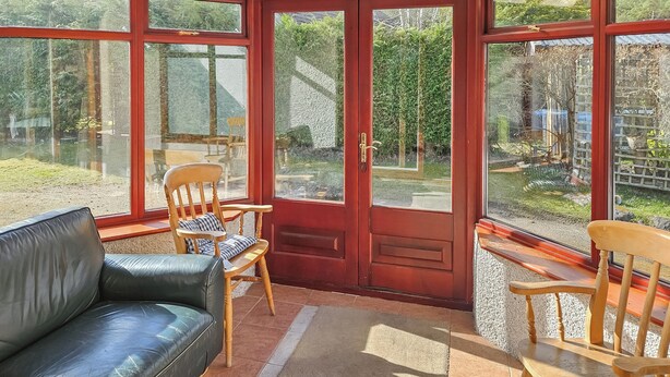 Une pièce baignée de lumière, avec une porte vitrée à cadre rouge, un canapé en cuir noir et deux fauteuils en bois dotés de coussins.