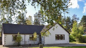 Une maison de plain-pied avec un toit recouvert de tuiles sombres, des murs extérieurs blancs et un petit jardin devant, agrémenté d’un banc en pierre.