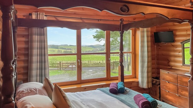 A wooden cabin room with a bed, a TV, and a window with a view of the countryside.