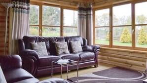 A log cabin interior with a leather sofa, a glass coffee table, and large windows offering a view of the outdoors.
