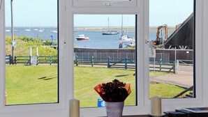 A view through a window with a grassy area, a flower arrangement, and a marina with boats and a crane in the background. - 4 bedroom accommodation in Wells-next-the-Sea (Wells-next-the-Sea)
