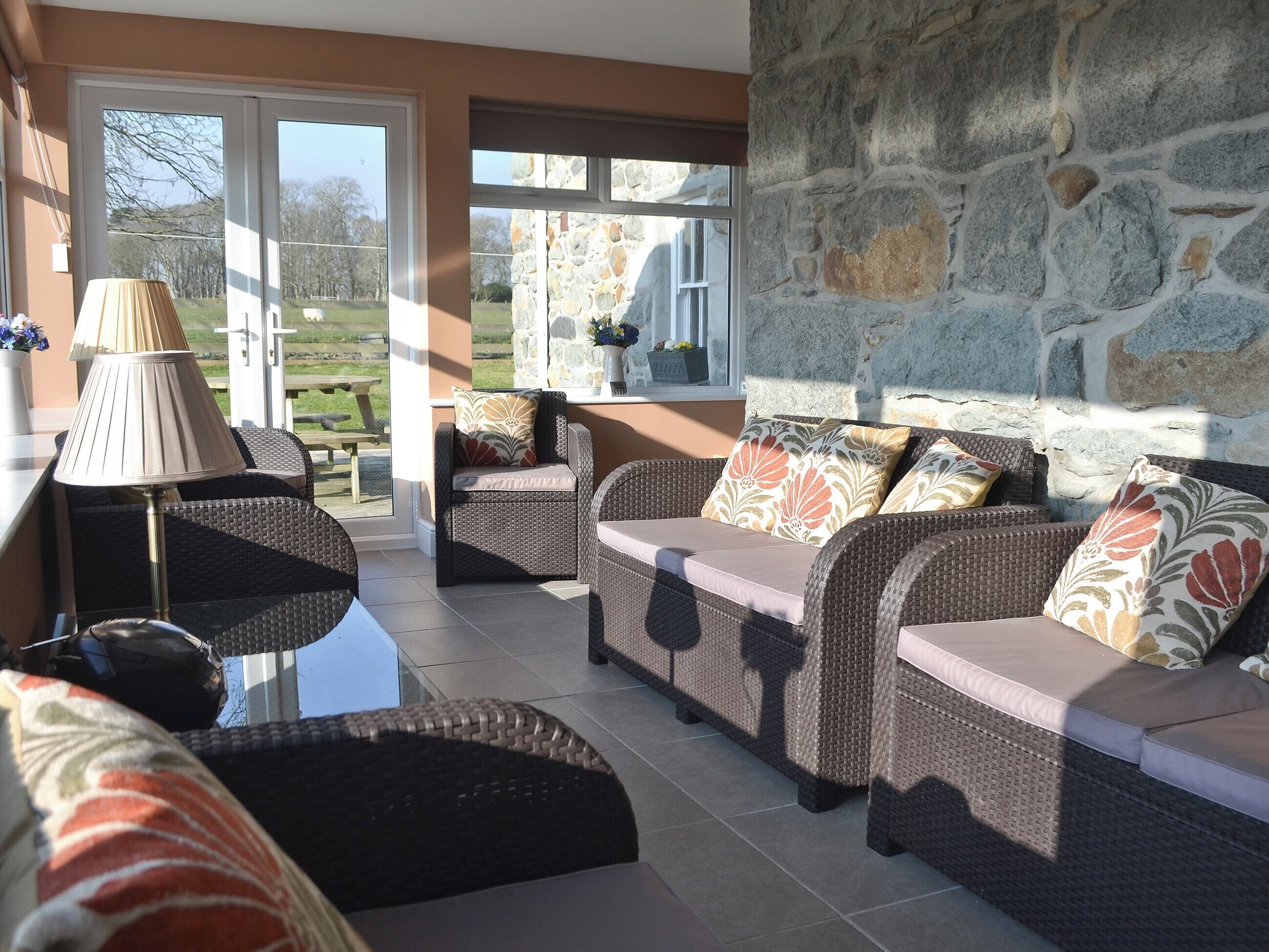 A sunlit room with wicker furniture, stone walls, and a view of a garden.