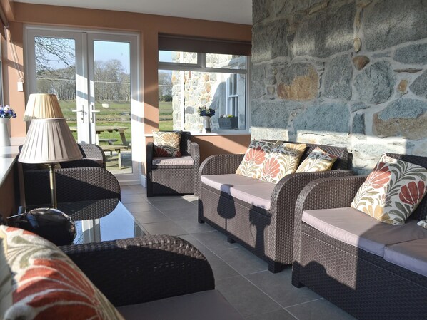 A sunlit room with wicker furniture, stone walls, and a view of a garden.